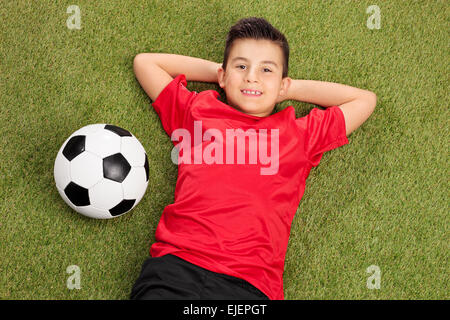 Carefree little boy in un rosso football jersey giacente su erba con una sfera accanto a lui Foto Stock