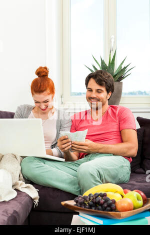 Close up molto felice coppia giovane con Laptop seduta sul lettino e gettare denaro euro nell'aria. Foto Stock
