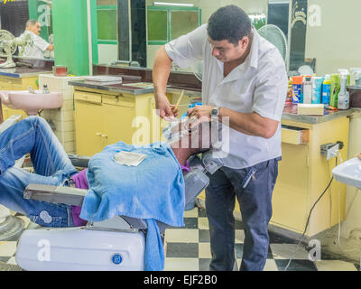 Uomo cubano si stende nel barbiere di sedia e ottiene la rasatura in un barbiere dal barbiere in Santiago de Cuba. Foto Stock