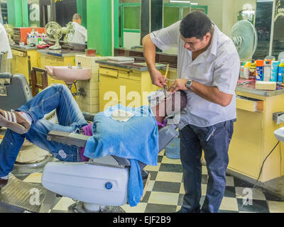 Uomo cubano si stende nel barbiere di sedia e ottiene la rasatura in un barbiere dal barbiere in Santiago de Cuba. Foto Stock