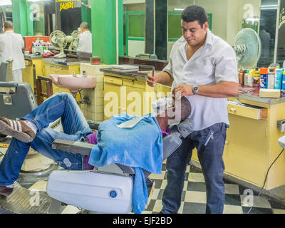 Uomo cubano si stende nel barbiere di sedia e ottiene la rasatura in un barbiere dal barbiere in Santiago de Cuba. Foto Stock