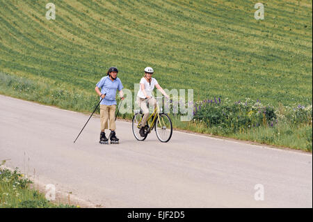 Coppia senior vigorosamente esercizio. L'uomo è nordico pattinaggio inline e la donna è in bicicletta. Si trova su una strada di campagna Foto Stock