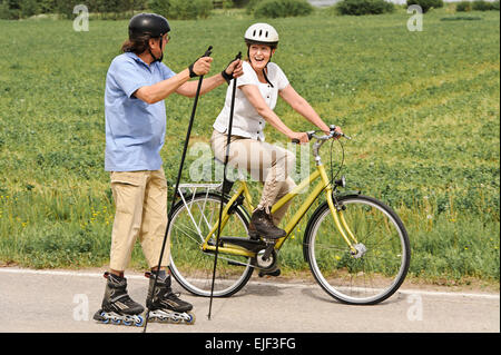 Coppia senior vigorosamente esercizio. L'uomo è nordico pattinaggio inline e la donna è in bicicletta. Si trova su una strada di campagna Foto Stock