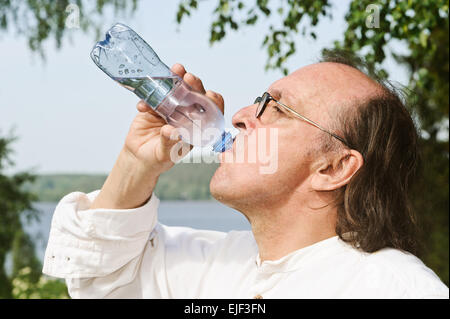 Sete senior uomo beve l'acqua da una bottiglia all'esterno. C'è un lago e la foresta in background. Foto Stock