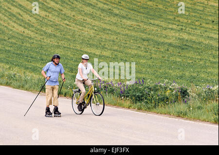 Coppia senior vigorosamente esercizio. L'uomo è nordico pattinaggio inline e la donna è in bicicletta. Si trova su una strada di campagna throu Foto Stock