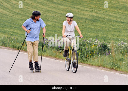 Coppia senior vigorosamente esercizio. L'uomo è nordico pattinaggio inline e la donna è in bicicletta. Si trova su una strada di campagna throu Foto Stock