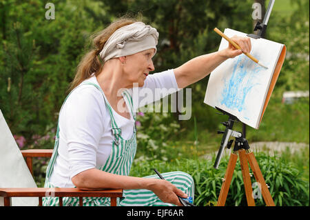Senior donna pittura all'esterno. Si siede in una sedia di tela di fronte al cavalletto. Il giorno è coperto dando una luce morbida Foto Stock