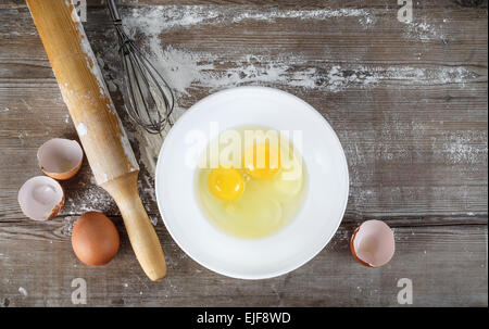 La cottura di sfondo con uova, uova crude in un piatto da portata, gusci d'uovo, la farina, il mattarello e frusta su un tavolo. Sfondo di legno. Top vie Foto Stock