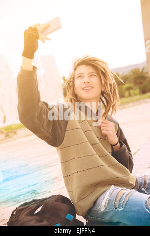 Ritratto di giovane ragazzo outdoor con capelli rasta sorridente con smart phone in un concetto di stile di vita con un caldo filtro applicato Foto Stock