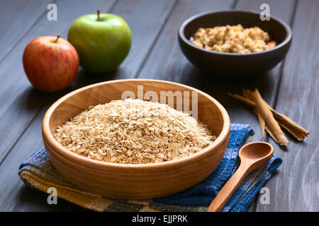 Materie di fiocchi d'avena in ciotola di legno con mele, bastoncini di cannella e una ciotola di frutta sgretolarsi in retro, fotografato in legno scuro Foto Stock