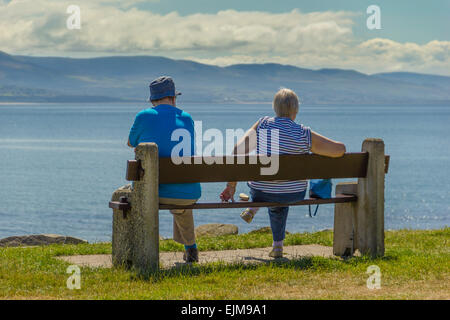 Coppia di anziani a prendere il sole su una panchina si affaccia su Cardigan Bay a Criccieth con Snowdonia Mountain Range in background. Foto Stock