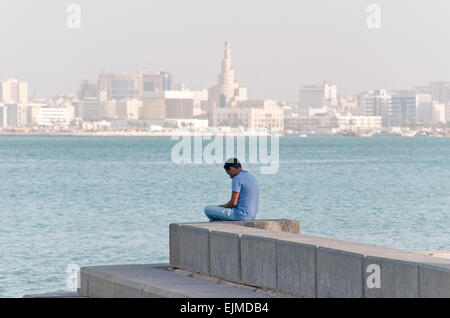 Un uomo si siede da solo a Doha, in Qatar, con il paesaggio e il Golfo Arabico in background. Foto Stock