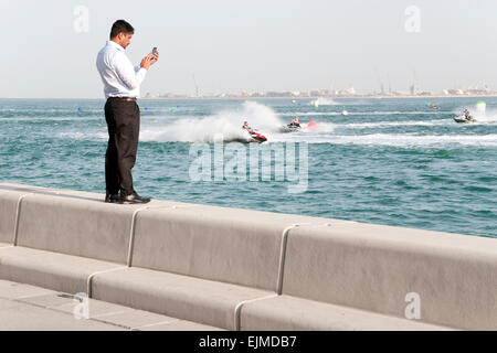Un uomo si erge sul lungomare di prendere un telefono cellulare immagine un jet-ski la concorrenza a Doha, in Qatar. Foto Stock
