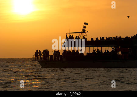 Un gruppo turistico di persone è la vela lungo l'acqua oceanica con un brillante tramonto in background. Foto Stock