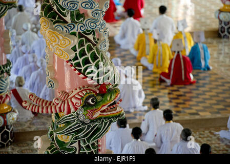 Caodaist discepoli seduto accanto a colonne colorate con draghi durante la cerimonia, Cao Dai temple, Tay Ninh Santa Sede, Tay Ninh, provincia di Tay Ninh, Vietnam, Dicembre Foto Stock