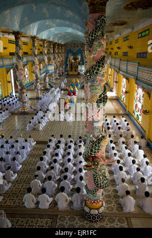 Caodaist discepoli seduto accanto a colonne colorate con draghi durante la cerimonia, Cao Dai temple, Tay Ninh Santa Sede, Tay Ninh, provincia di Tay Ninh, Vietnam, Dicembre Foto Stock