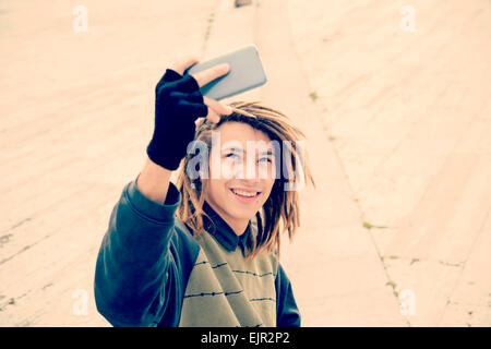 Ritratto di giovane ragazzo outdoor con capelli rasta sorridente con smart phone in un concetto di stile di vita con un caldo filtro applicato Foto Stock