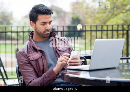 Uomo indiano mediante telefono cellulare e computer portatile a tavola all'aperto Foto Stock