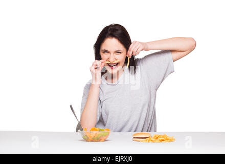 Naturale donna espressiva giocando con patatine fritte, avendo di fronte junk e cibo sano, isolato su bianco Foto Stock