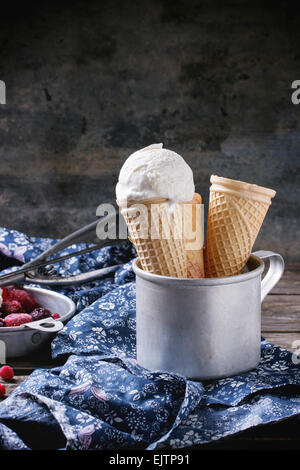 Gelato alla vaniglia in coni e svuotare i coni di waffer, servita nel recipiente di metallo con frutti di bosco surgelati e cucchiaio su un tavolo di legno wi Foto Stock
