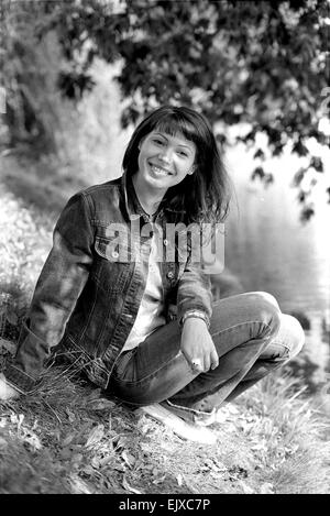 Giovane donna sorridente seduti in riva al lago sotto l'albero. Foto Stock