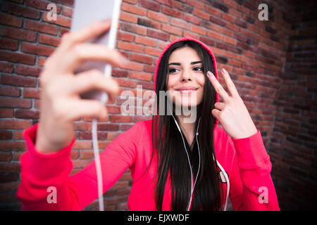 Felice bella donna rendendo selfie foto sullo smartphone. Indossare in panno sportive. In piedi su un muro di mattoni Foto Stock