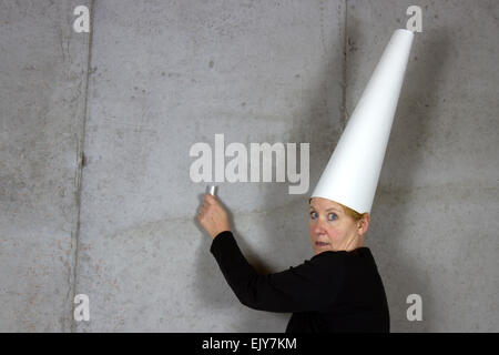 Donna che indossa un cappello dunce con chalk rivolta verso una parete di cemento Foto Stock