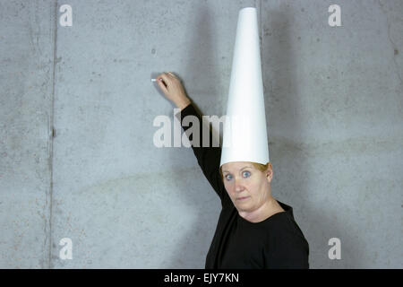 Donna che indossa un cappello dunce usando il gesso per scrivere sulla parete Foto Stock