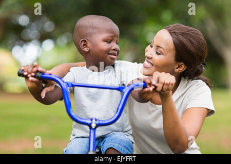 Piuttosto giovane donna africana aiutando il suo adorabile figlio un giro in bici Foto Stock