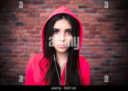 Bella giovane donna con le cuffie su un muro di mattoni. Guardando alla fotocamera. Indossare in rosa il cofano Foto Stock