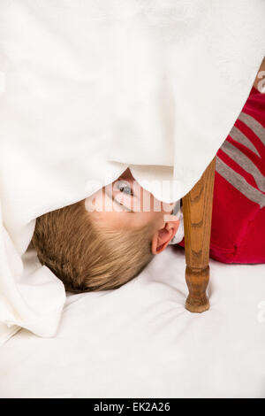 6 anno vecchio ragazzo scherzosamente che spuntavano da sotto un tavolo drappeggiato Foto Stock