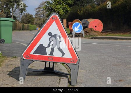 Un appaltatori roadworks cartello di avviso in una zona rurale strada residenziale. Mantenere la sinistra in segno e bobine di cavo in background Foto Stock