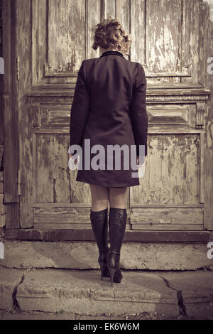 Vista posteriore della bella ragazza in un coatagainst weathered porta in città, solitudine, concetto di depressione Foto Stock