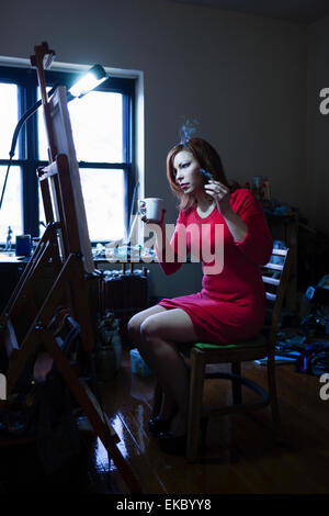 Metà donna adulta seduti nella parte anteriore del cavalletto, azienda di sigarette e di bevande calde Foto Stock