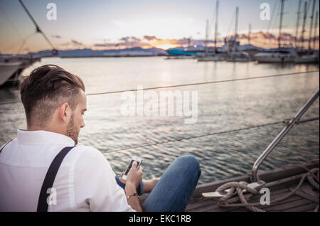 Giovane uomo utilizza lo smartphone su yacht, Cagliari, Sardegna, Italia Foto Stock