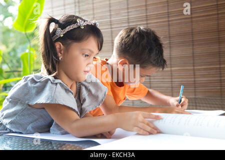 Coppa CuteÊlittle ragazza asiatica la lettura di un libro di storia seduto accanto a un fratello più anziano assorbiti in attività di colorazione in home ambientale Foto Stock