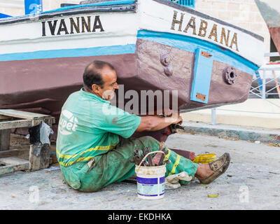 Una mezza età uomo cubano siede sulla terra come egli le riparazioni il propulsore sulla sua barca a l'Avana, Cuba. Foto Stock