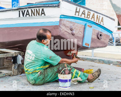 Una mezza età uomo cubano siede sulla terra come egli le riparazioni il propulsore sulla sua barca a l'Avana, Cuba. Foto Stock