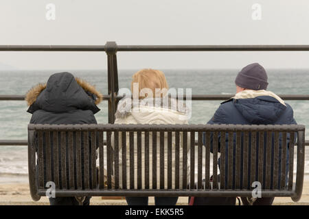 Tre turisti che si godono la loro vacanza sat su banco cercando in mare un freddo nuvoloso giorno di primavera in Looe, Cornwall, Inghilterra Foto Stock