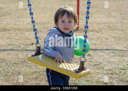 Un bambino di 2 anni boy tenendo una palla e in piedi dall'oscilla in un parco giochi. Foto Stock
