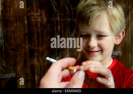 Ritratto di bambino biondo ragazzo Sigaretta fumare con legno scuro sfondo dietro di lui. Adult man mano che offre una sigaretta a boy Foto Stock