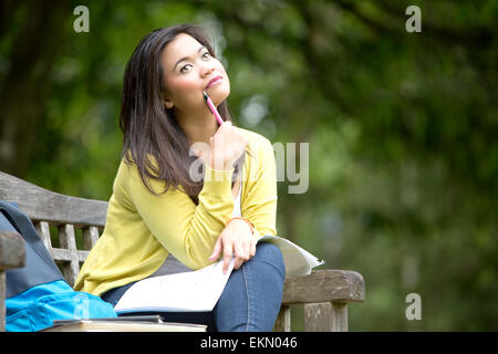 Bellissima giovane università asiatiche o studente di college seduta sul banco di legno nel parco, con libri e zaino a fianco, pensando w Foto Stock