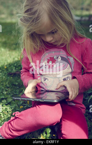 Bambino bella ragazza bionda seduta sul prato nel parco e gioca con la tavoletta digitale Foto Stock