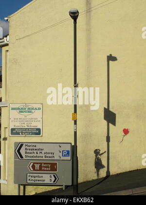 Un Banksy murale della ragazza con palloncino rosso sulla parete a Brixham Devon con ombre e segnaletica stradale e Berry Head Hotel segno Foto Stock