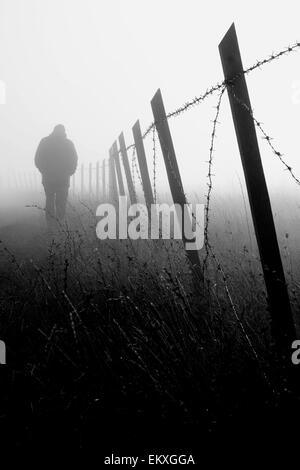 Uomo che cammina vicino al recinto di filo spinato nella fitta nebbia Foto Stock