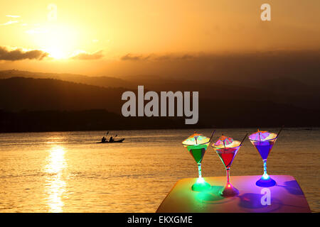 Tre bicchieri da cocktail illuminati a LED, di colore rosso, verde e blu, su un tavolo da spiaggia al tramonto. Per ulteriori informazioni, vedere la sezione "ulteriori informazioni" Foto Stock
