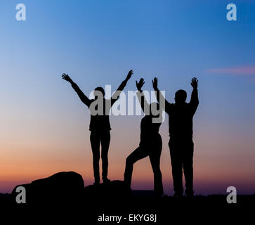 Silhouette di una famiglia felice con le braccia sollevate fino contro il bellissimo cielo colorati. Tramonto d'estate. Paesaggio Foto Stock