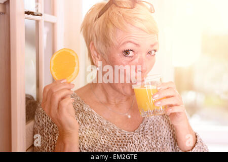 Chiudere alto Medioevo donna bionda di bere succo di arancia e di trattenimento di un arancione fetta di frutta mentre appoggiata sulla parete Foto Stock