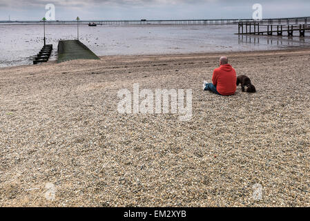 Un uomo e il suo cane seduto sulla spiaggia della città di Southend in un giorno nuvoloso. Foto Stock