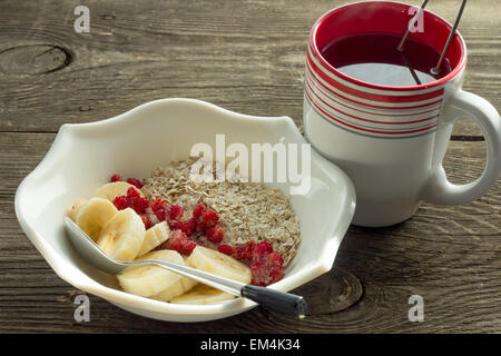 Farina di avena, di banana e le fragole congelate in bianco ciotola, cucchiaio e la tazza di tè sul tavolo rustico Foto Stock